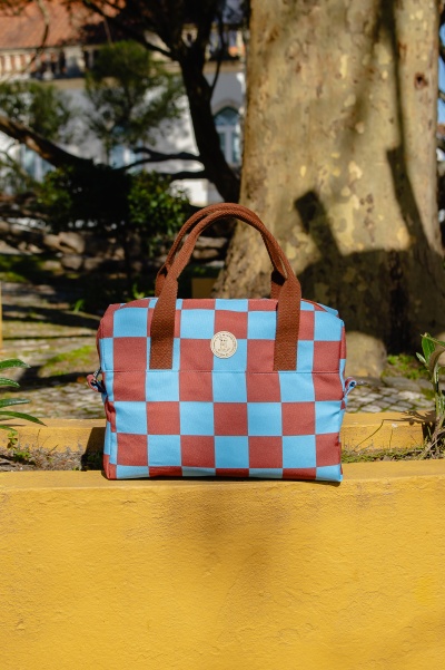Mala azul e castanha quadriculada com alças castanhas num muro amarelo com árvores atrás