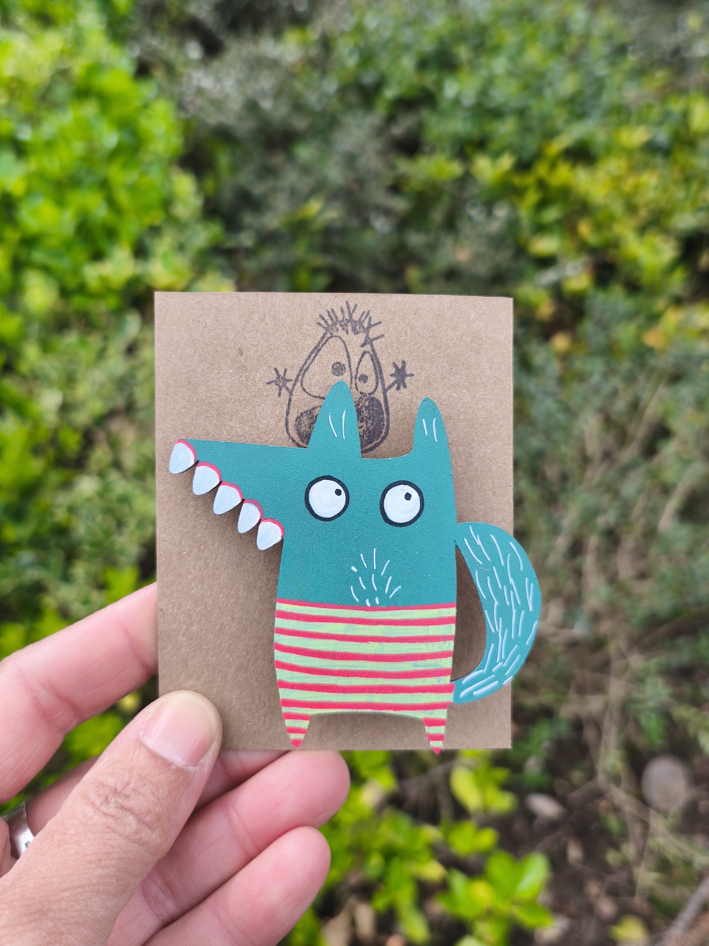 Alfinete lobo! pin artesanal de animal azul esverdeado com riscas amarelas e vermelhas segurado por mão