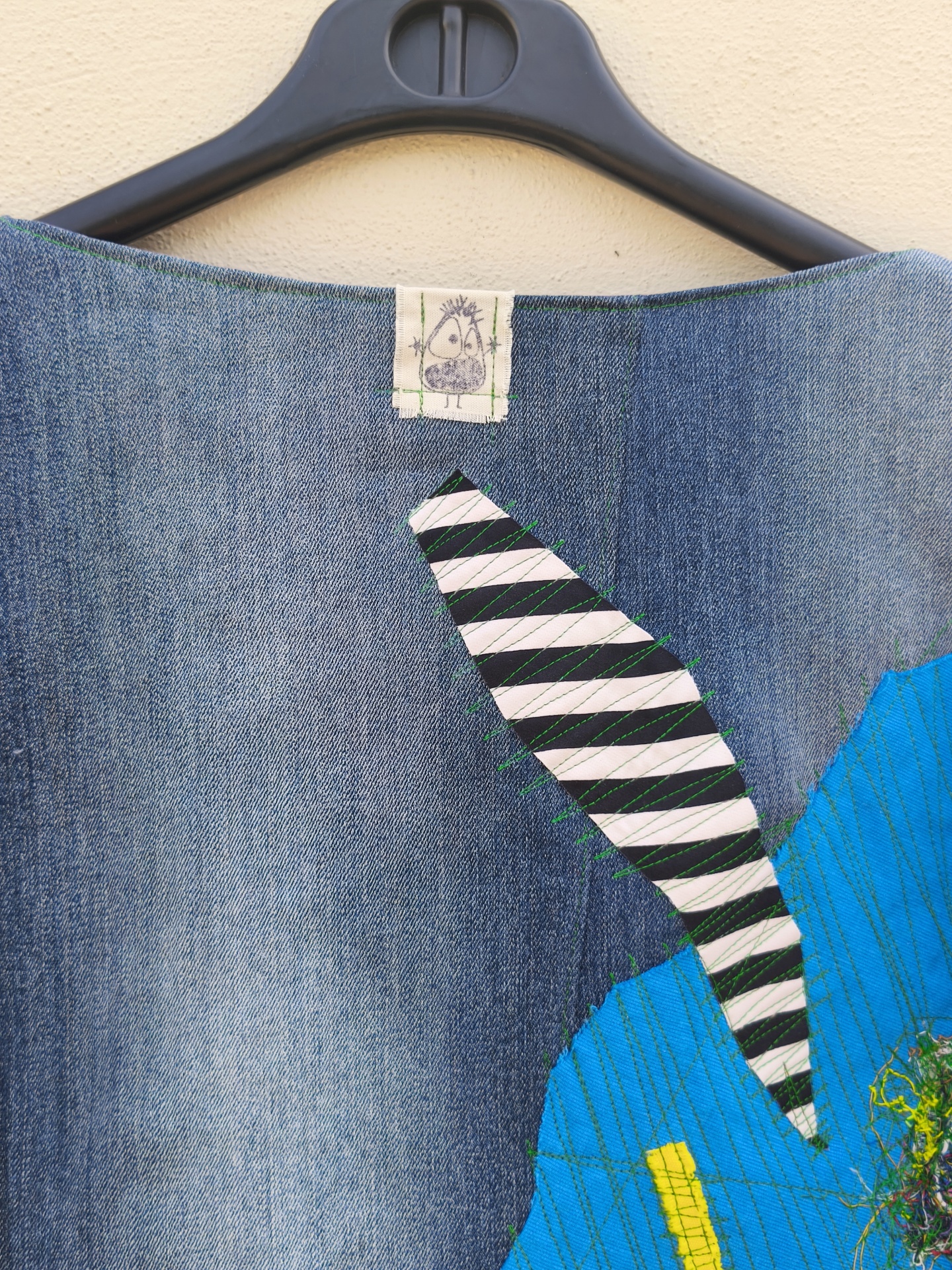 Casaco de ganga Trapo #4 Peça de roupa em ganga azul com aplicações de tecidos riscados e coloridos pendurada num cabide preto