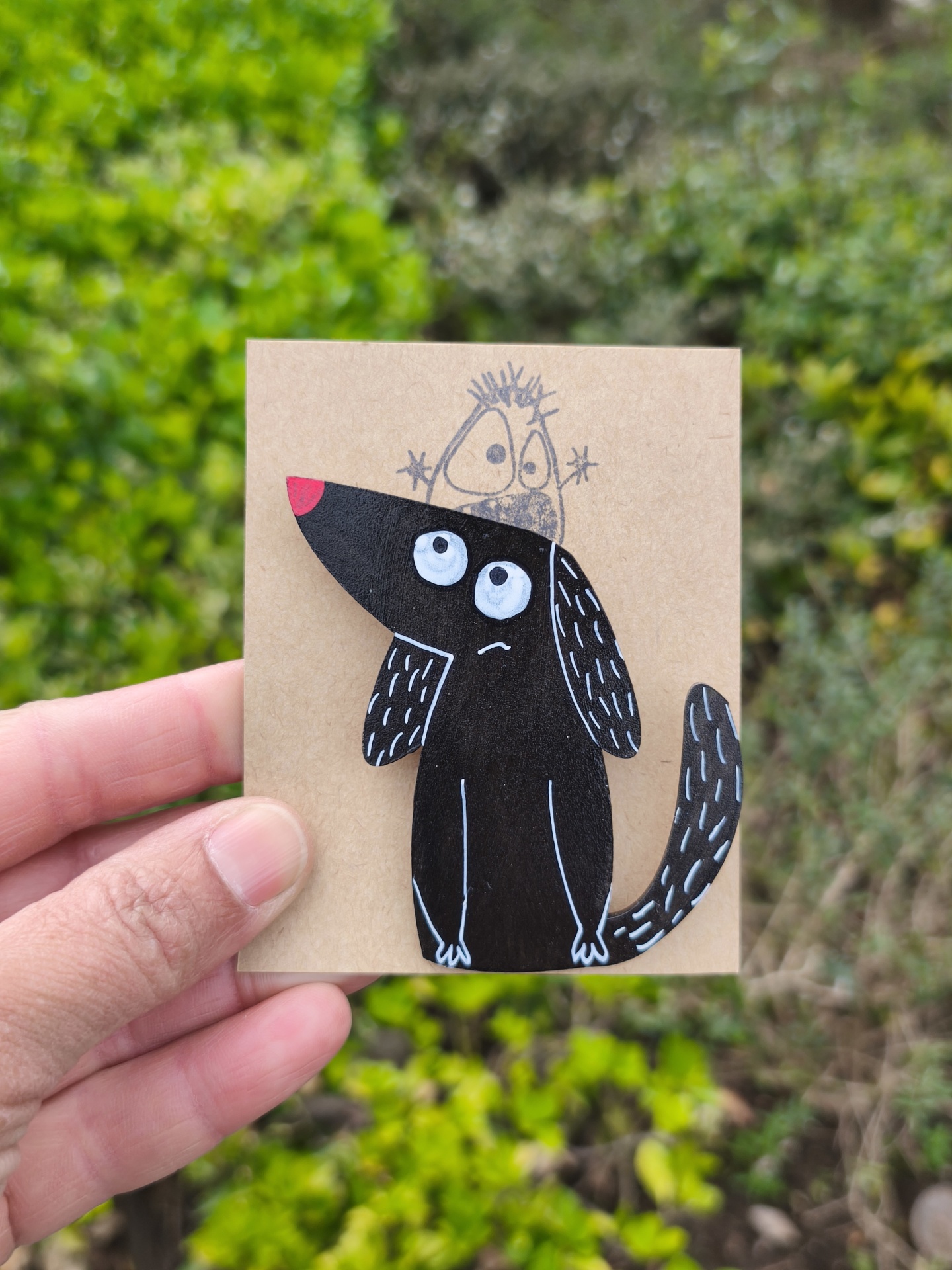 Alfinete cão! Broche de cão preto em madeira pintada montado num cartão castanho com ilustração desenhada ao fundo