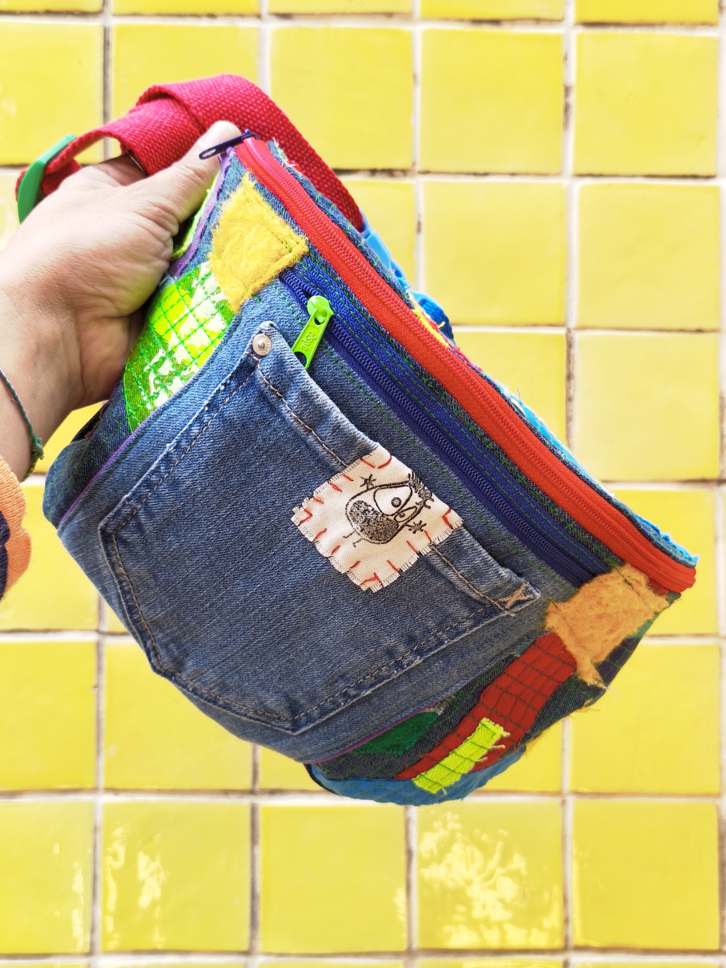 Bolsas de cintura Trapo! Mala colorida com bolso de ganga e vários remendos, segurada por uma mão ante fundo de azulejos amarelos.