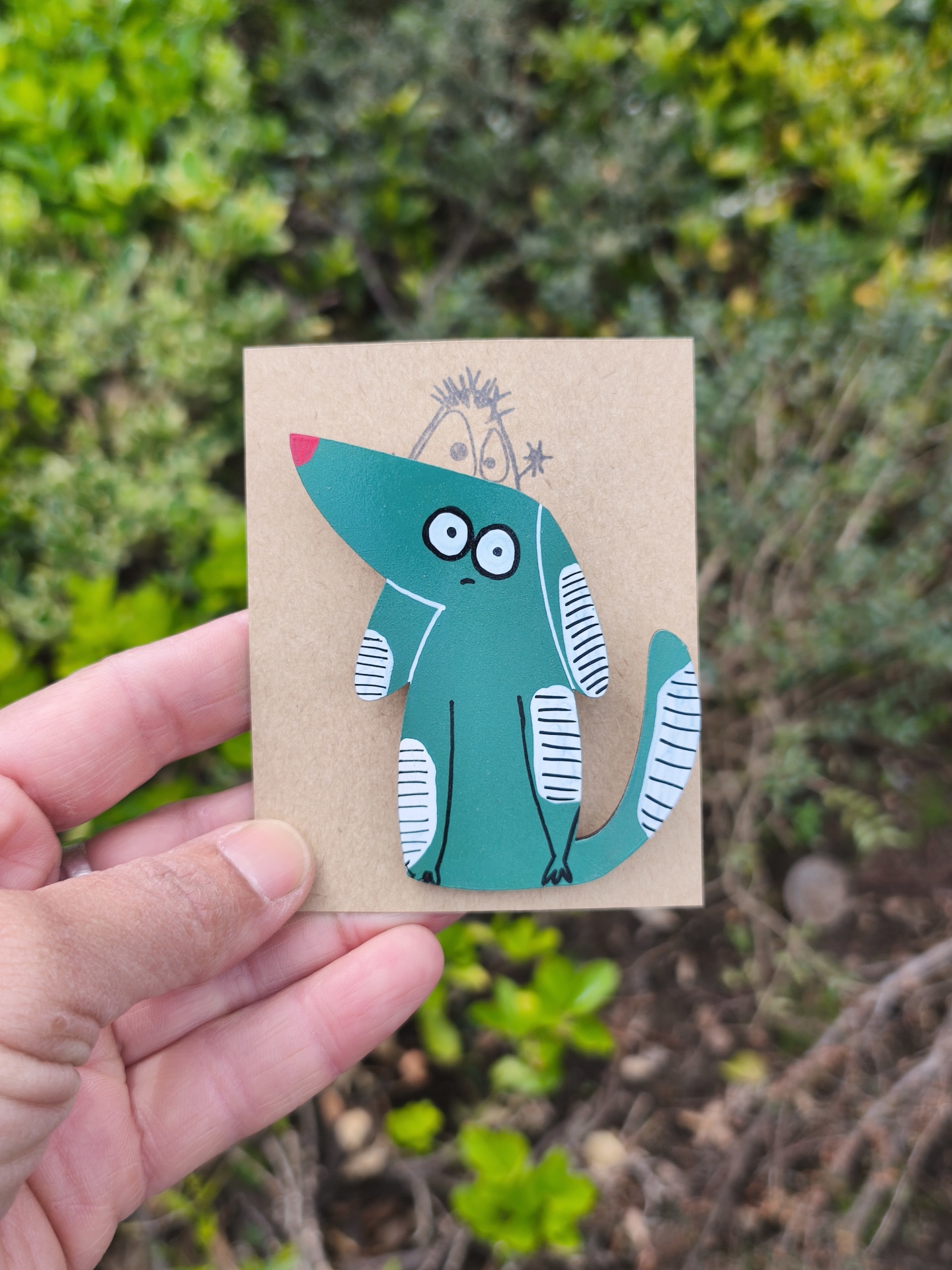 Alfinete cão! Broche de cão verde com manchas brancas preso a cartão castanho, mão a segurá-lo, fundo com plantas