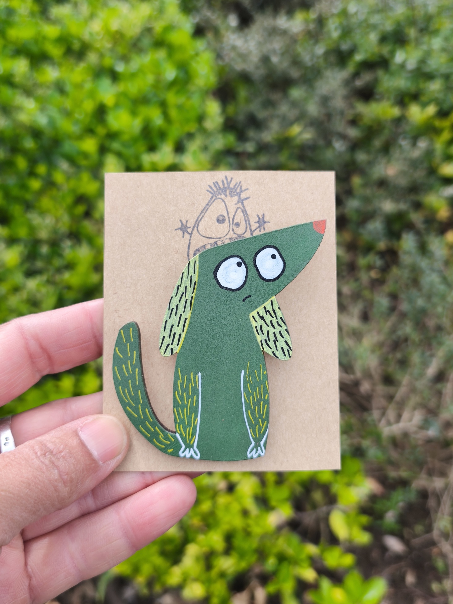 Alfinete cão! Broche/pendente em forma de cão verde com olhos grandes e nariz vermelho, sobre cartão castanho, mão segurando, fundo com plantas verdes
