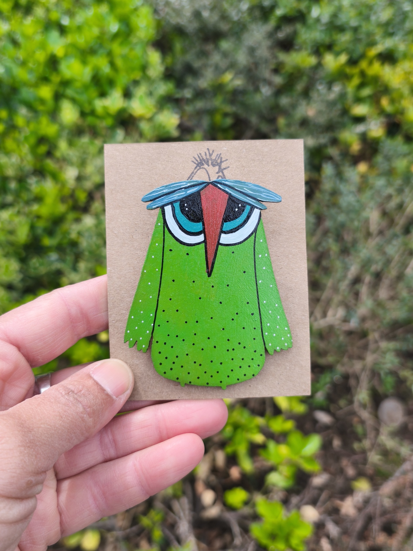 Alfinete mocho! Pin de madeira em forma de pássaro verde com olhos azuis e bico vermelho, em cartão castanho sendo segurado à mão