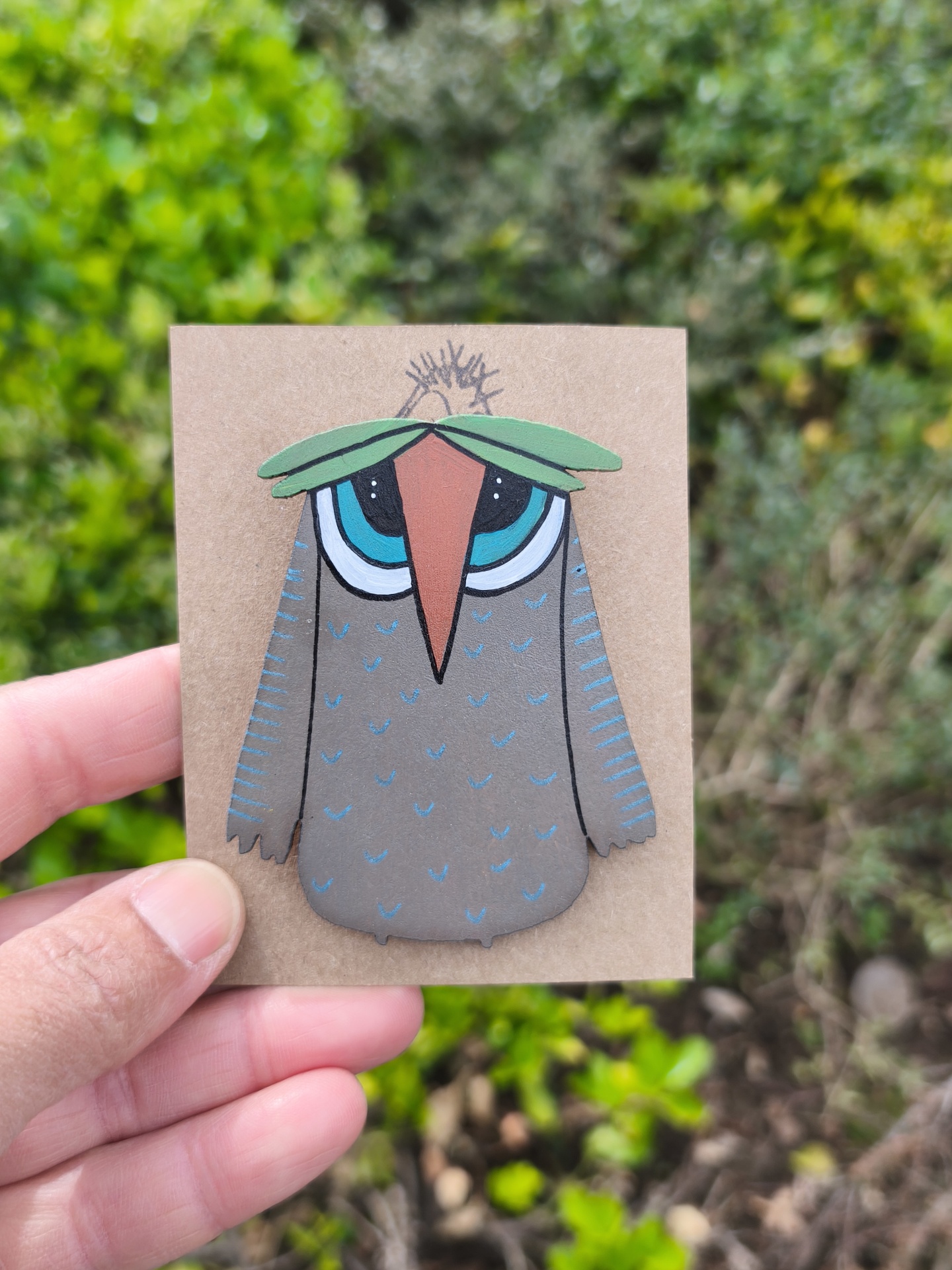 Alfinete mocho! Broche de coruja estilizada em madeira segurado por mão, com fundo de folhagem verde