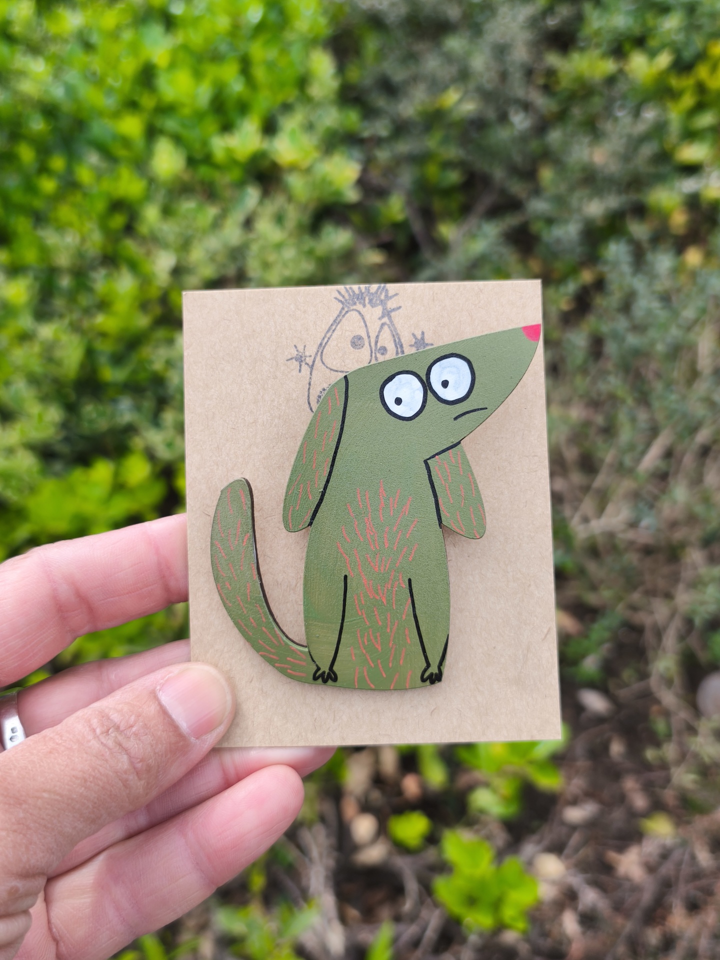 Alfinete cão! Broche em madeira pintada de cão verde com olhos grandes, em cartão castanho, com fundo de plantas outdoors