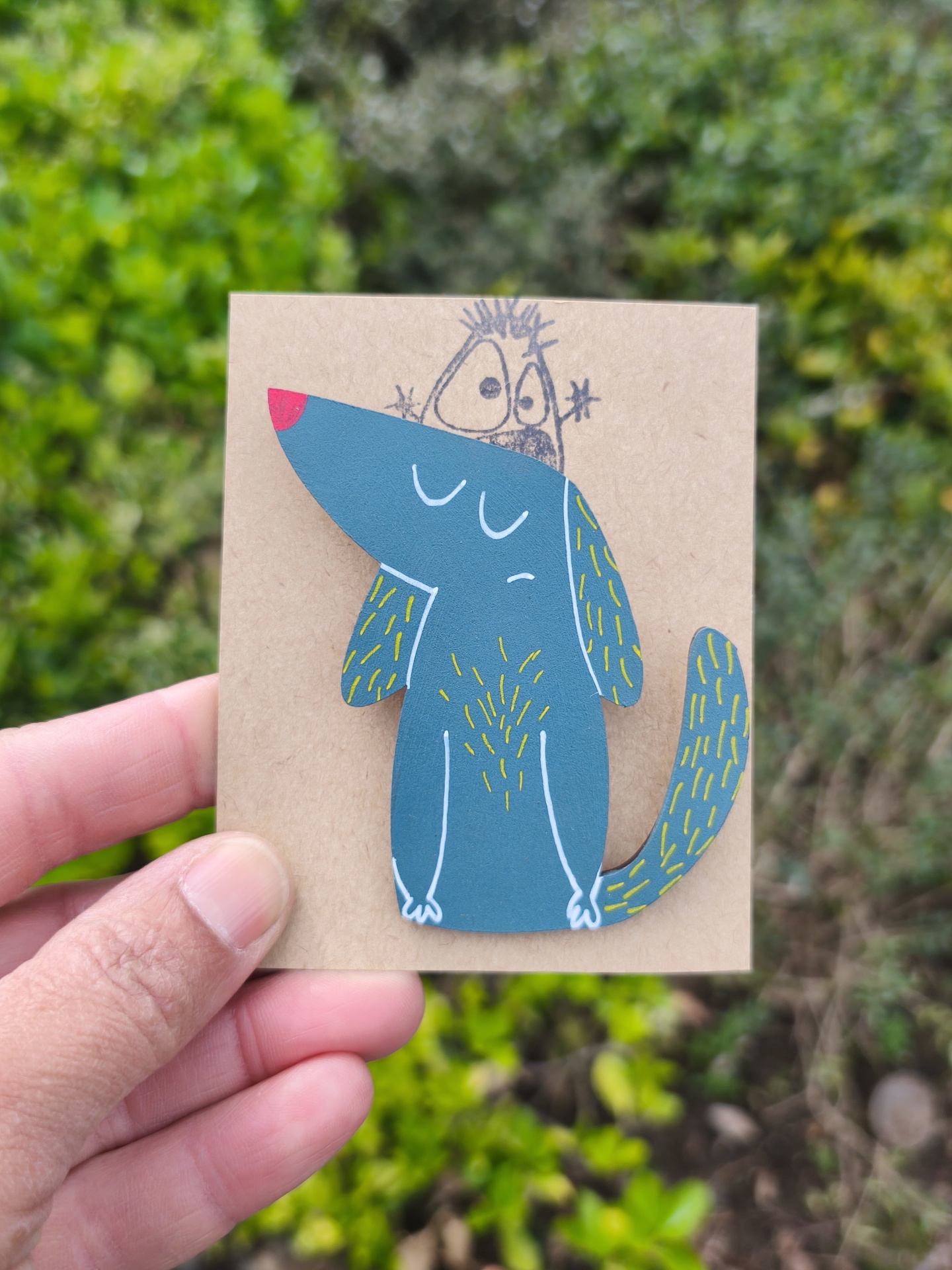 Alfinete cão! Cartão decorativo com ilustração de cão azul, segurado com fundo de plantas verdes.