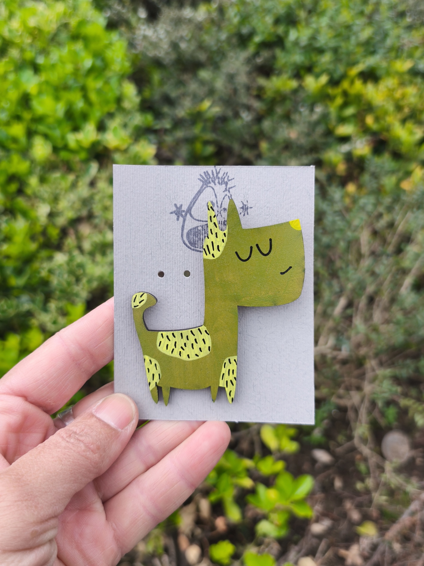 Alfinete cão #2! Broche de madeira em forma de cão verde com manchas amarelas, em cartão cinzento com desenho a tinta preta, segurado por mão com fundo de plantas.