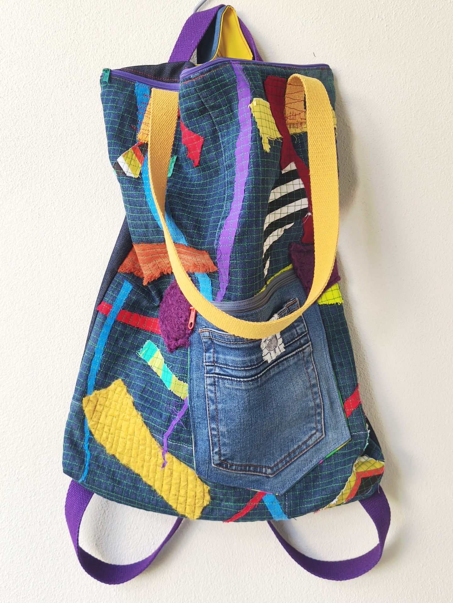 Bolsa/Mochila Mochila colorida patchwork com bolso jeans e alças roxas e amarelas pendurada na parede