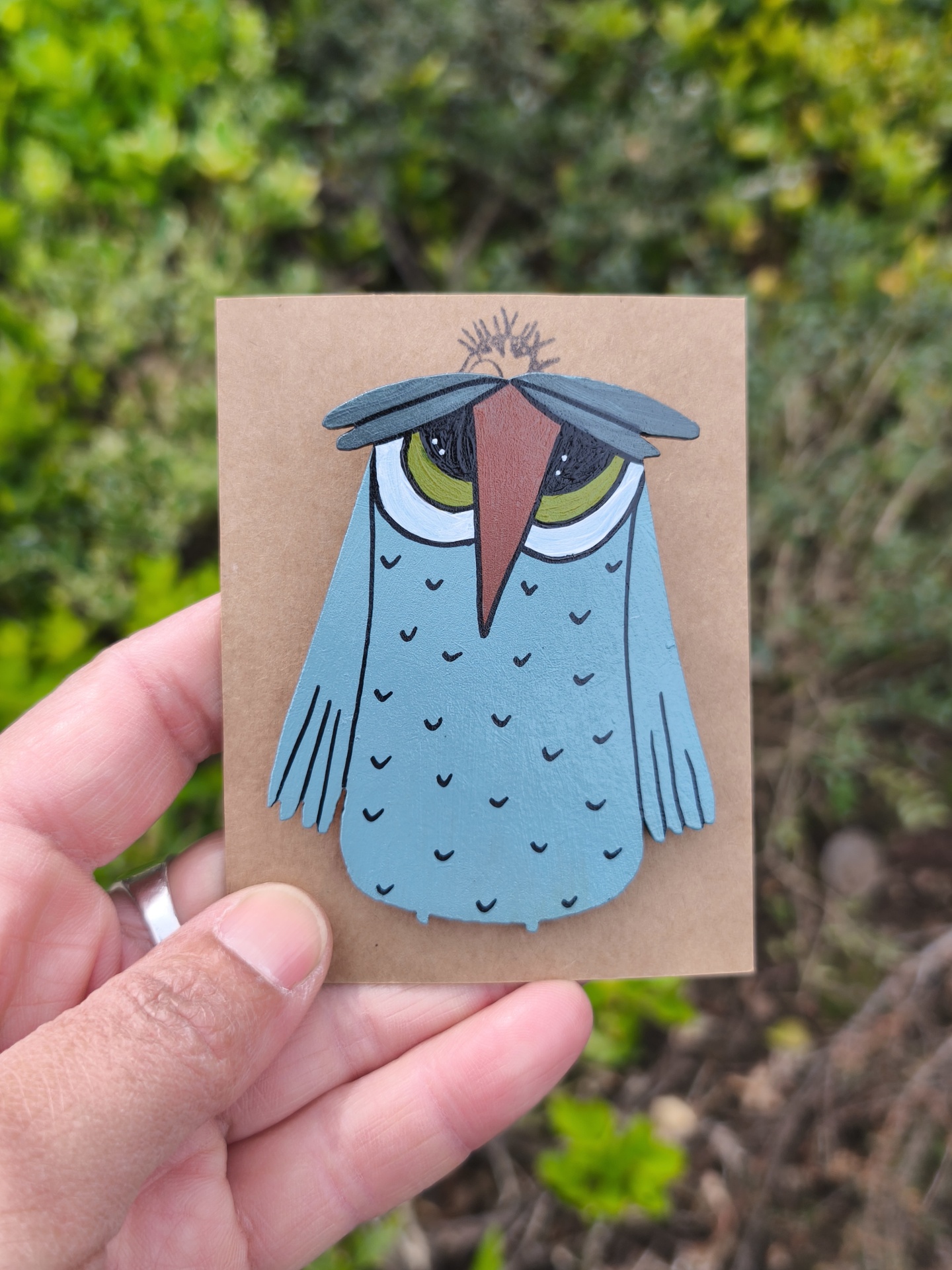 Alfinete mocho! Broche de coruja azul em cartão castanho com fundo verde desfocado