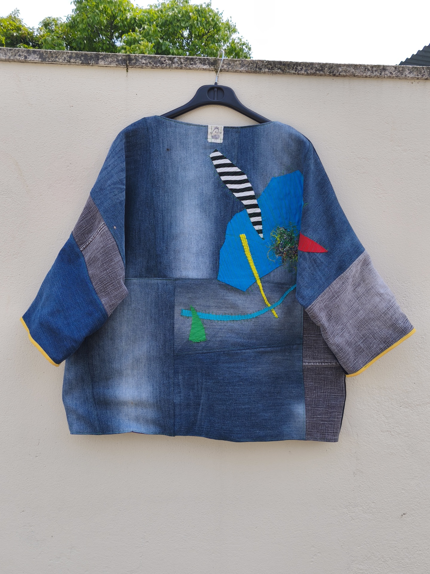 Casaco de ganga Trapo #4 Blusa de tecido azul com retalhos coloridos pendurada numa parede branca