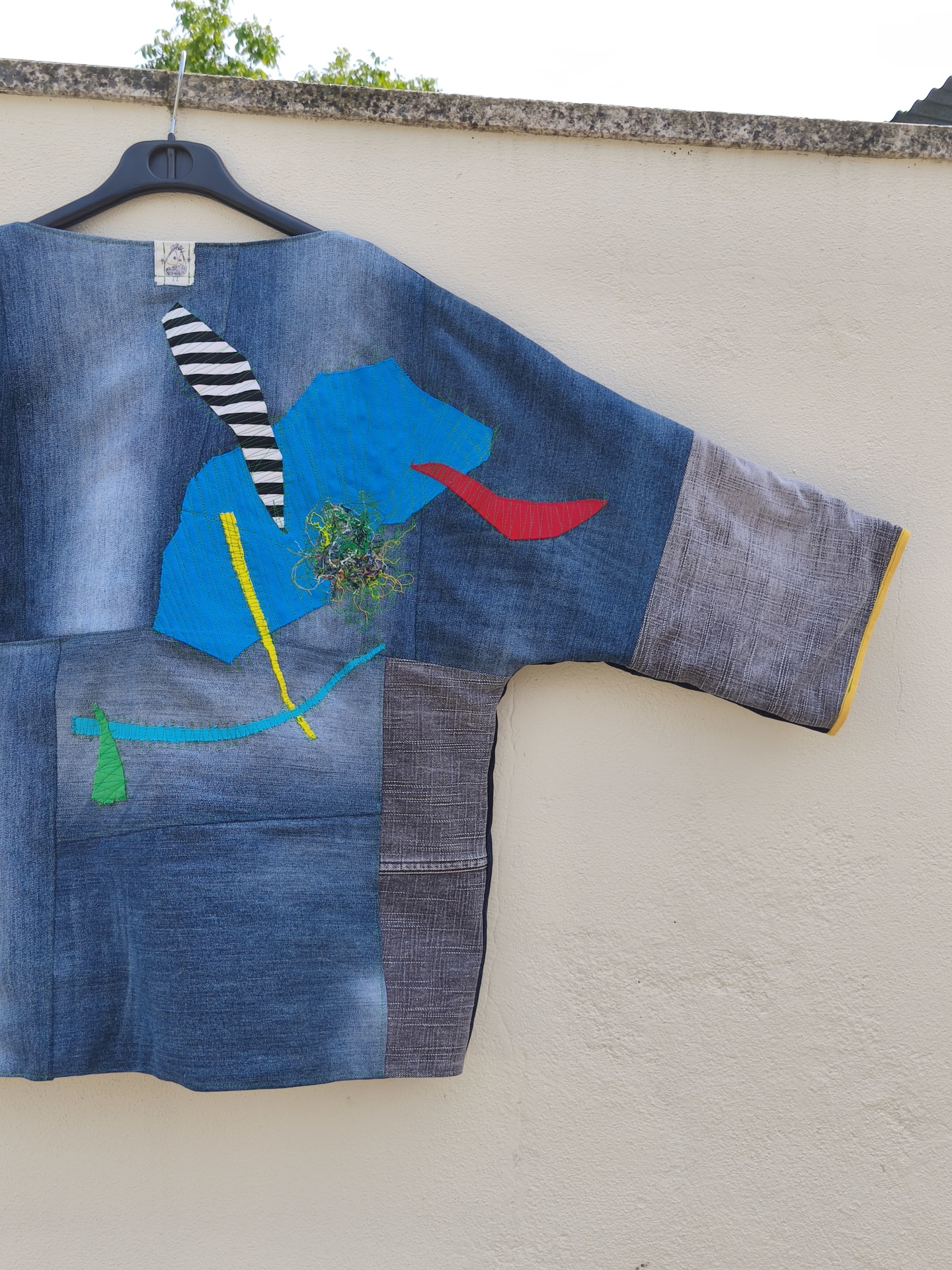 Casaco de ganga Trapo #4 Túnica jeans azul e cinzento com aplicações coloridas pendurada numa cabide preta