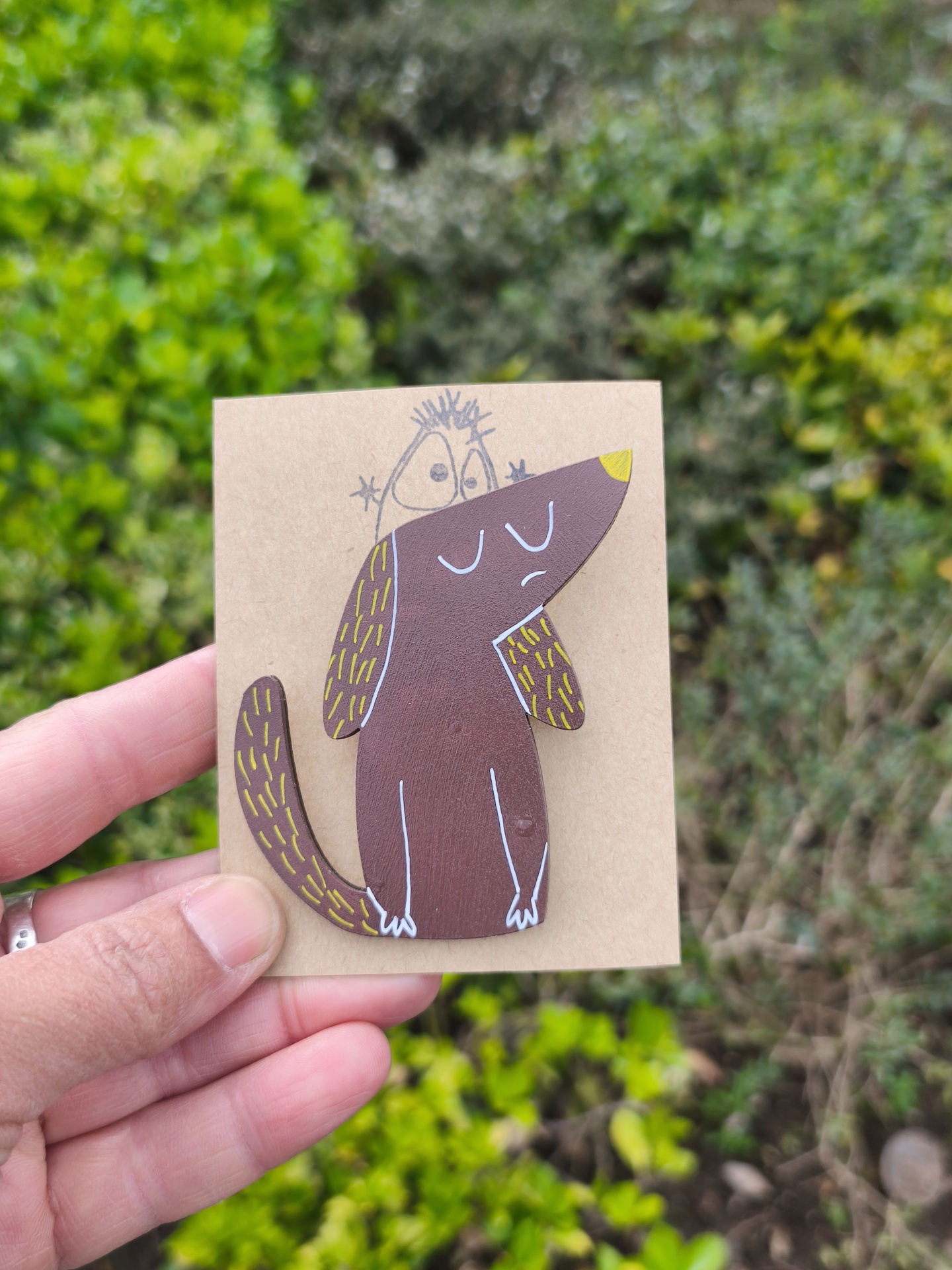 Alfinete cão! Broche castanho com desenho de cão em cartão castanho contra fundo de vegetação verde