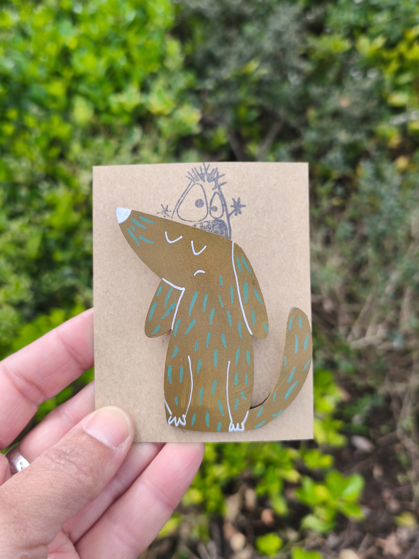 Alfinete cão! Cartão com ilustração de cão castanho e personagem desenhada, segurado por mão, com fundo de folhas verdes.