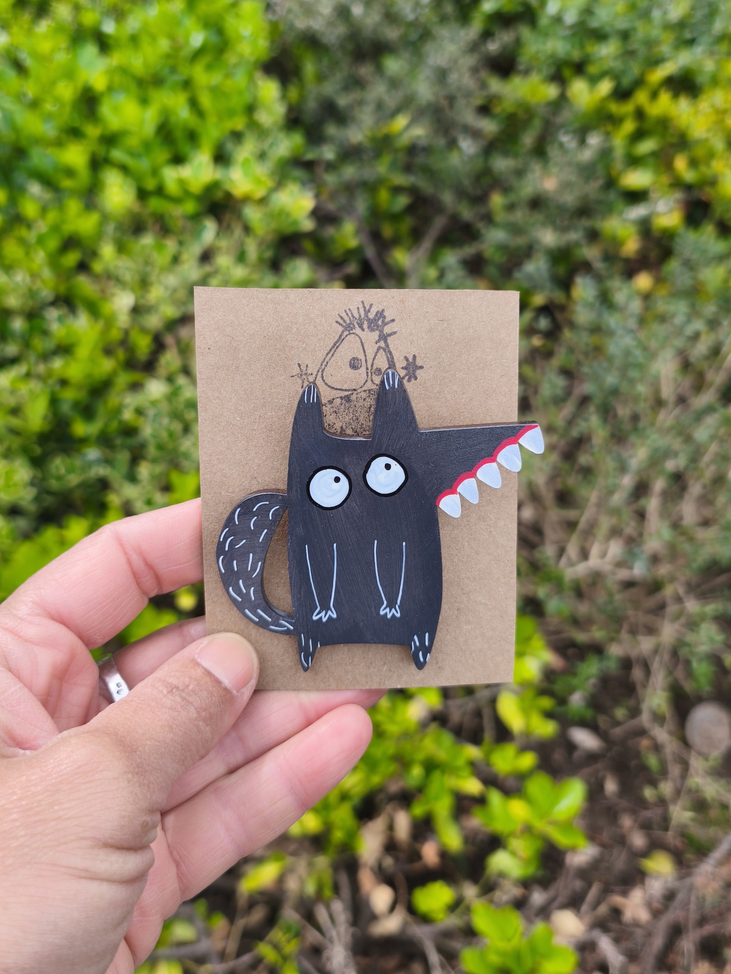 Alfinete lobo! Broche em madeira pintado à mão com figura de animal preto e cauda cinzenta, olhos grandes, dentes brancos e vermelhos, sobre cartão castanho.