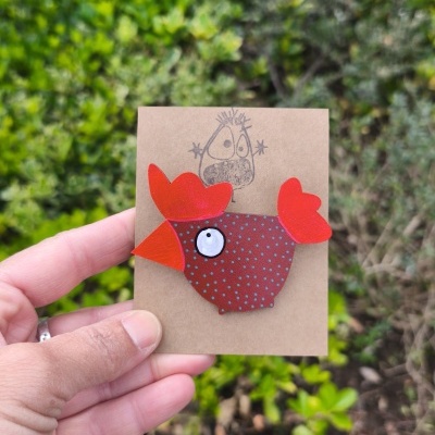 Broche em forma de galinha castanha e vermelha em cartão castanho com desenho preto segurado por uma mão.