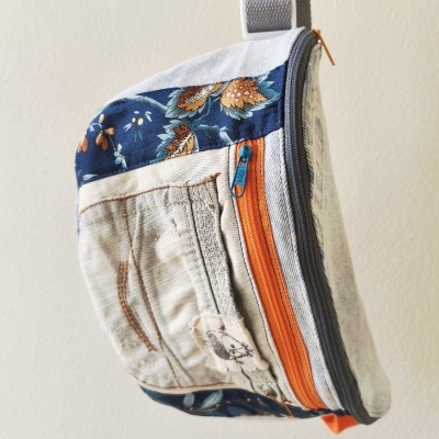 Mala tipo sling com tecido claro, detalhes florais azuis e zíperes laranja e cinza
