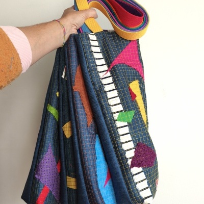 Mala de tecido azul com padrão colorido e alça multicolorida sendo segurada por uma mão.