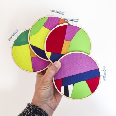 Quatro bastidores de bordado coloridos com padrões geométricos sendo segurados na mão