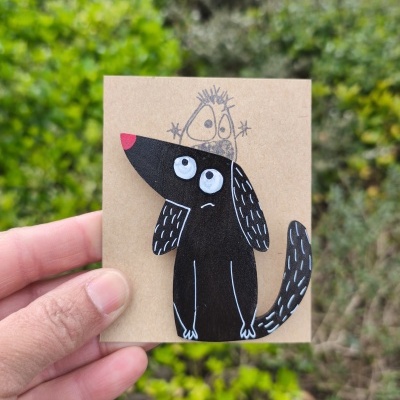Broche de cão preto em madeira pintada montado num cartão castanho com ilustração desenhada ao fundo
