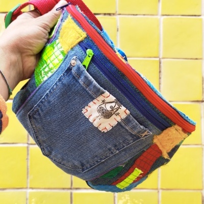 Mala colorida com bolso de ganga e vários remendos, segurada por uma mão ante fundo de azulejos amarelos.