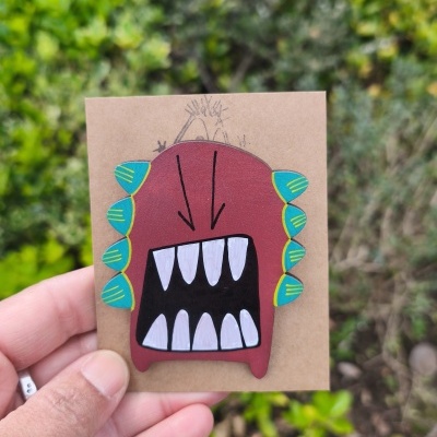 Broche de monstro colorido em madeira pintada, com dentes brancos e detalhes azuis e amarelos