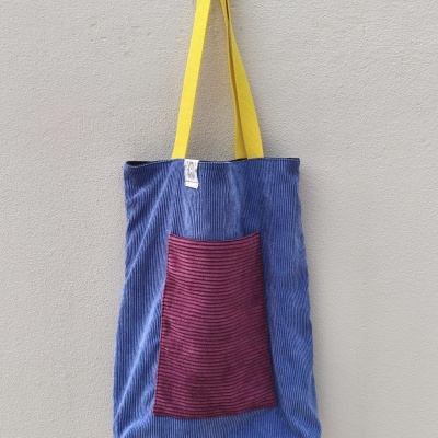 Mala tote azul com bolso vermelho e alças amarelas pendurada numa parede