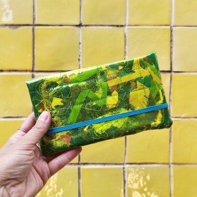 Carteira verde e amarela com elástico azul segurada por mão com fundo de azulejos amarelos