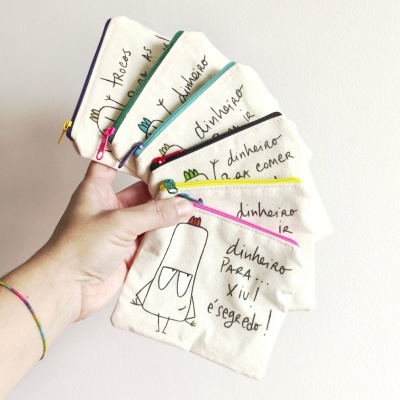 Carteiras pequenas de tecido bege com fechos coloridos e desenhos a caneta, seguradas por uma mão.