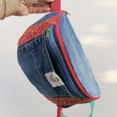 Riñonera de mezclilla azul con detalles rojos y cremallera roja, mostrando etiqueta textil y asa para colgar