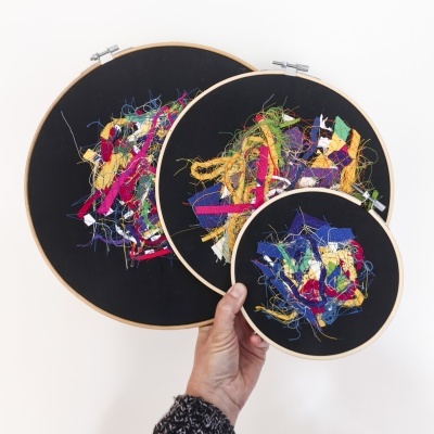 Três bastidores redondos com bordados abstratos coloridos em tecido preto.