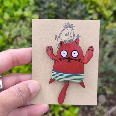 Broche de madeira com figura de gato vermelho e saia azul e amarela em base castanha ao ar livre