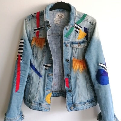 Casaco de ganga azul claro com bordados coloridos e texturas felpudas pendurado numa cabide preta