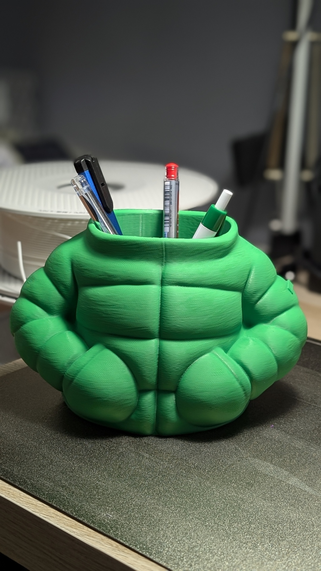 Suporte para canetas verde em forma de tronco musculado sobre mesa.