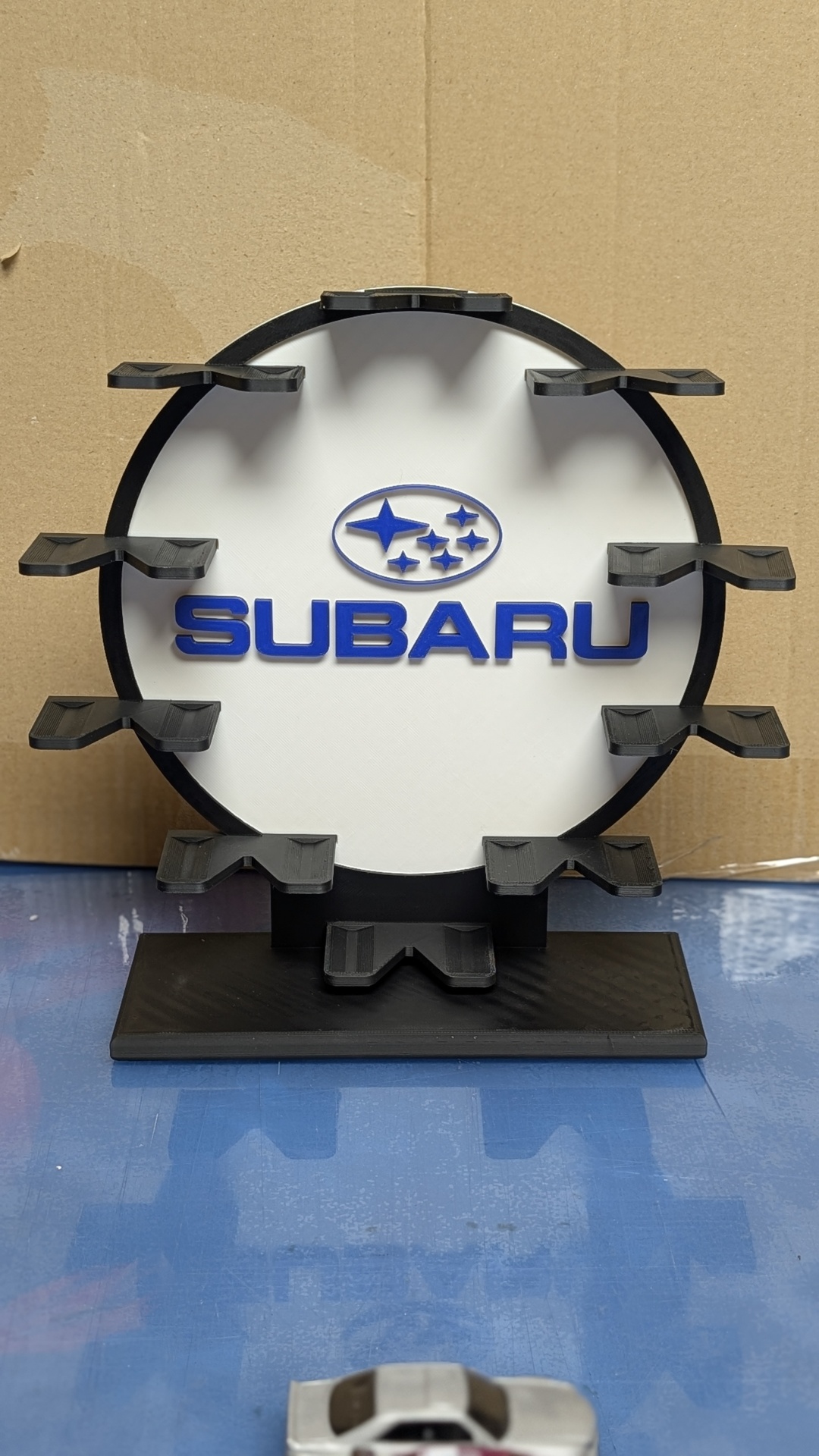 Suporte circular com logótipo da Subaru azul sobre fundo branco e base preta.
