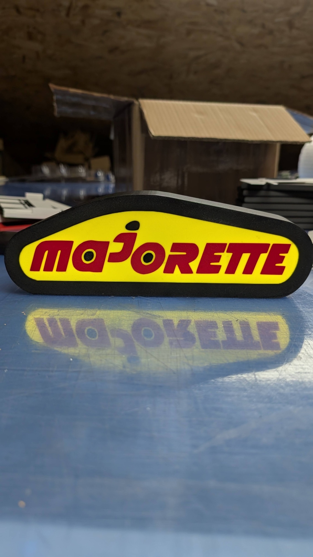 Logótipo Majorette amarelo e vermelho com borda preta sobre superfície azul