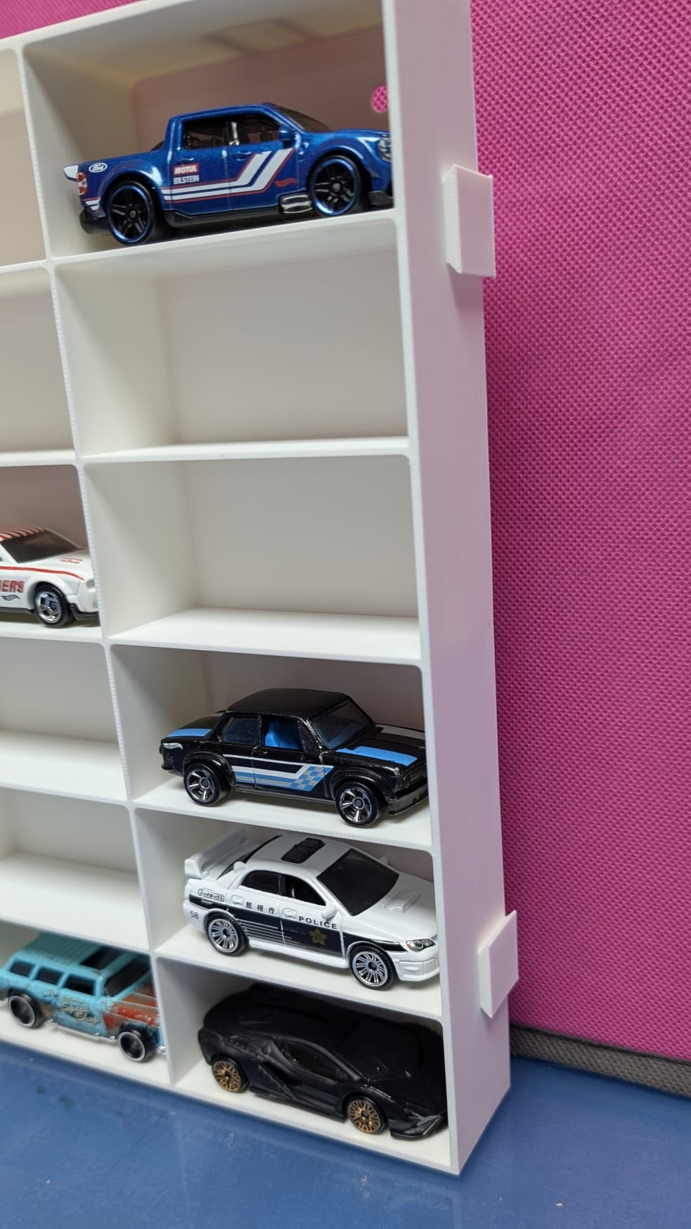 Carrinhos de brinquedo em miniatura em estante branca ao lado de parede cor-de-rosa.