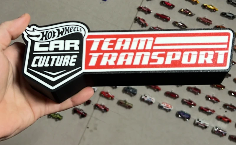 Objeto decorativo Hot Wheels com texto Team Transport e miniaturas de carros ao fundo