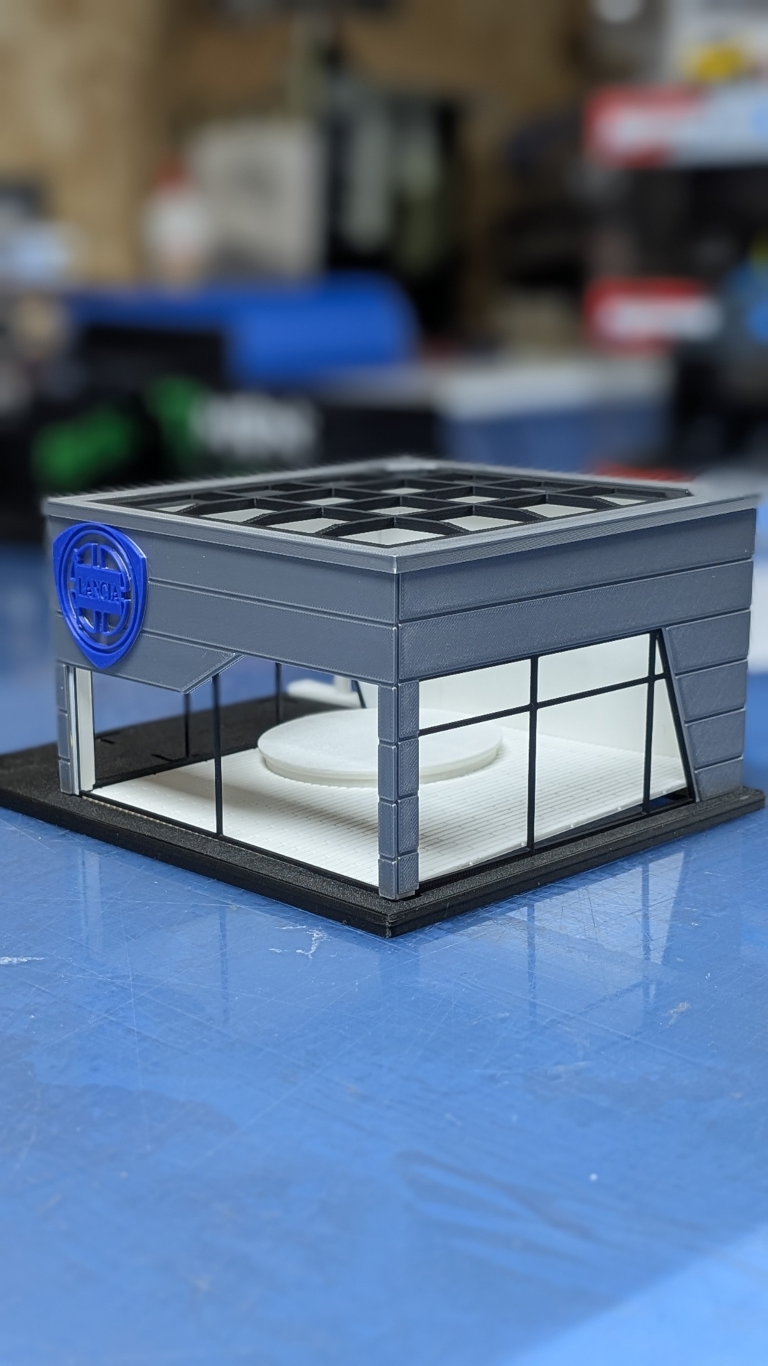 Maqueta de edifício com estrutura cinzenta e janelas de vidro, logotipo azul LANCIA