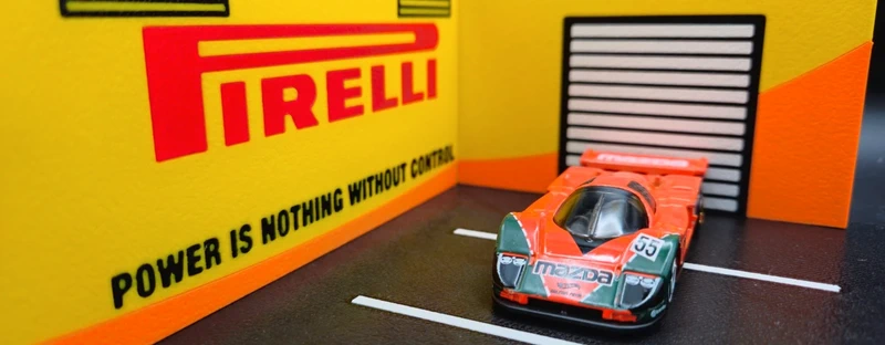 Modelo miniatura de carro de corrida Mazda com cores vermelho, verde e branco e número 55 numa garagem com marca Pirelli.