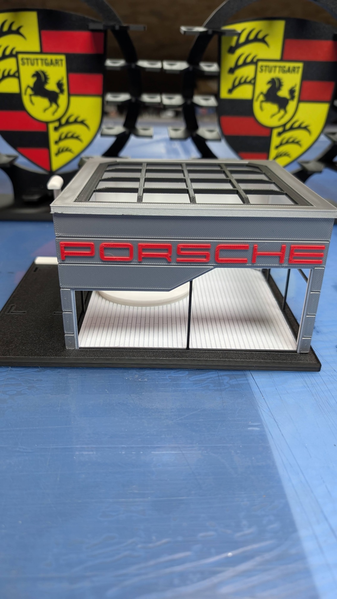 Miniatura de garagem Porsche com letras vermelhas, estrutura cinzenta, chão branco riscado e escudos Porsche ao fundo