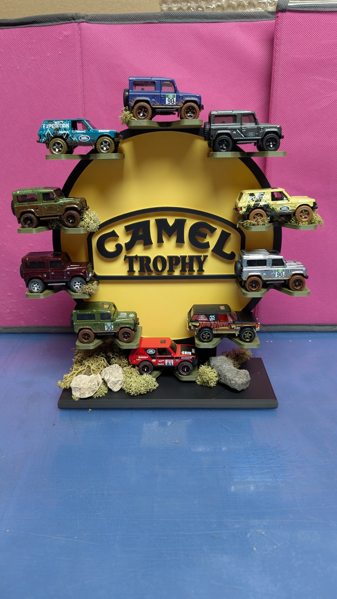 Miniaturas de carros Land Rover exibidas em suporte CAMEL TROPHY com decoração natural.