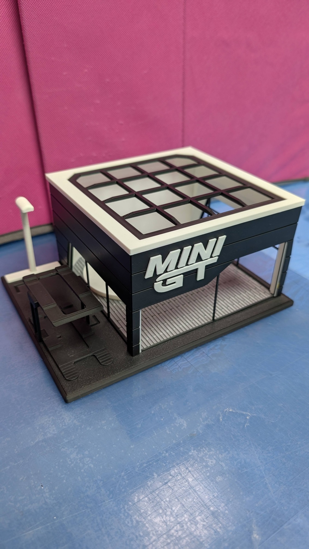 Miniatura loja Mini GT em plástico preto e branco sobre superfície azul com fundo rosa