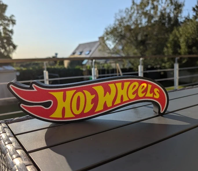 Placa decorativa Hot Wheels em madeira com texto amarelo sobre fundo vermelho em mesa exterior