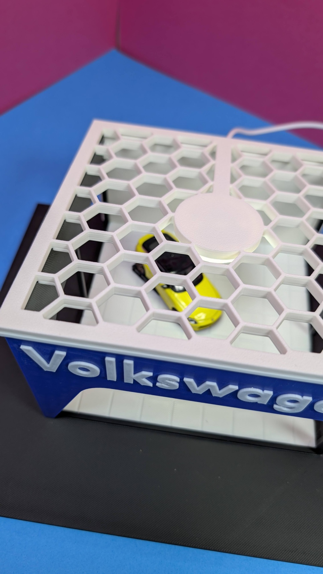 Miniatura de carro amarelo dentro de estrutura branca e azul com padrão hexagonal e texto VOLKSWAGEN.