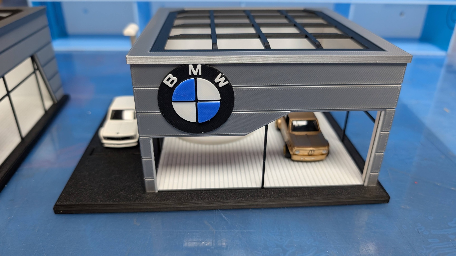 Miniatura de concessionária BMW cinza com dois carros em exposição e logótipo BMW frontal