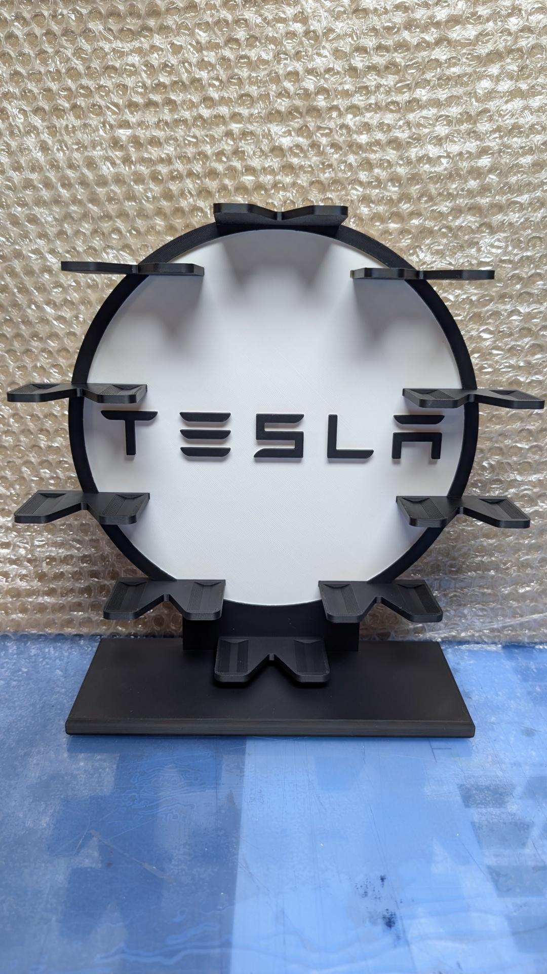 Suporte redondo preto e branco com texto Tesla e prateleiras em fundo de plástico bolha