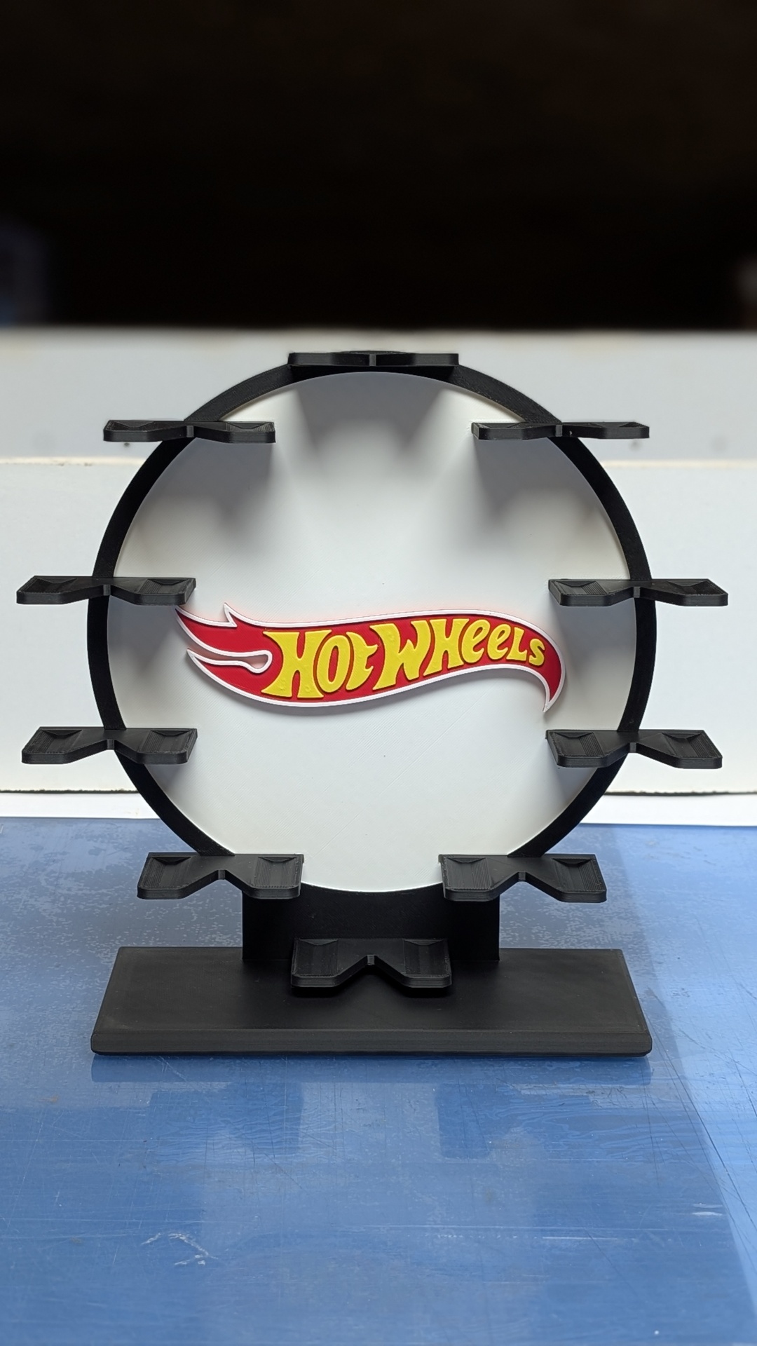 Expositor preto circular para carros Hot Wheels com logo ao centro sobre superfície azul