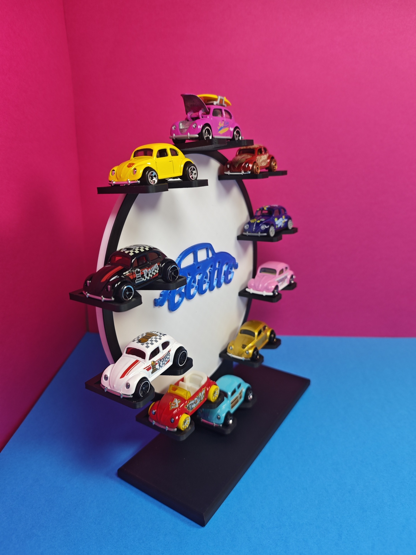 Miniaturas de carros Volkswagen Beetle em expositor preto e branco, com texto Beetle azul no centro, sobre fundo rosa e base azul.