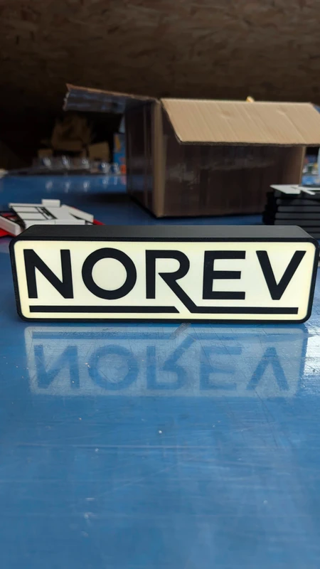 Placa branca com texto preto NOREV sobre superfície azul e caixa ao fundo